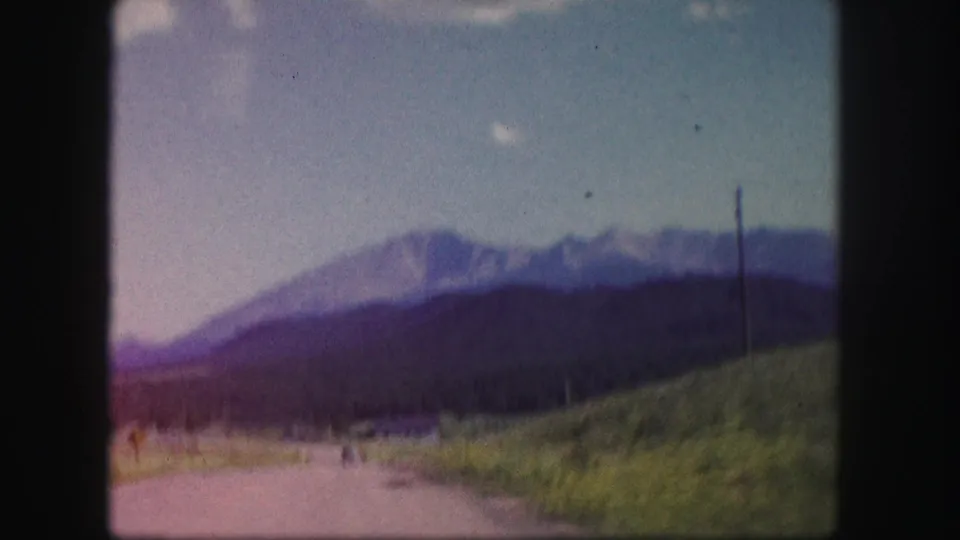Soft 1958 Colorado travel frame showing a road edge, grassy slope, utility pole, forested foothills, and distant mountains.