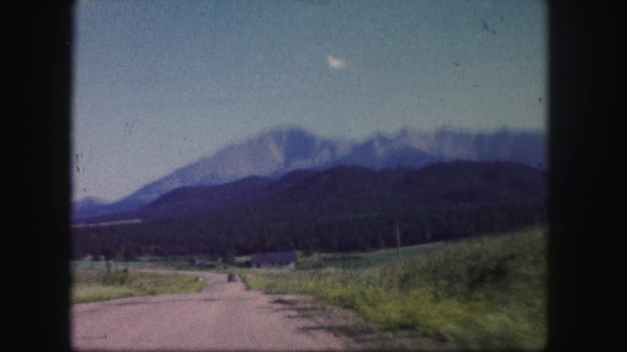 1958 Colorado 8mm frame showing a rural dirt road, grassy fields, tree line, and hazy mountain peaks in the distance.