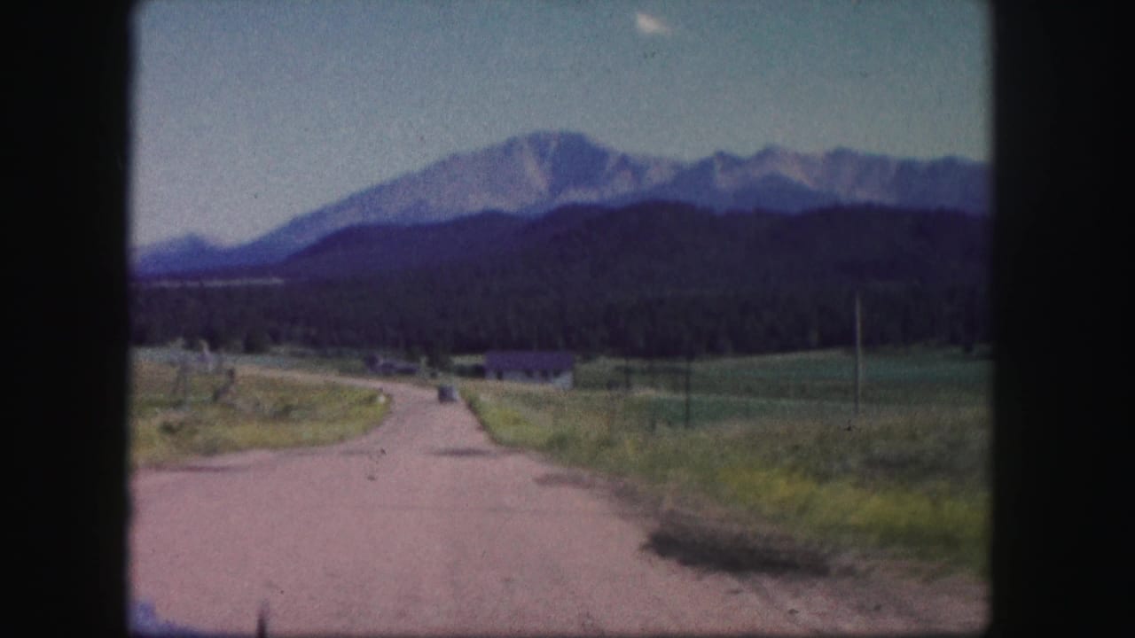 1958 Colorado home-movie frame with an isolated road, fields, utility poles, a small distant structure, and mountain peaks beyond.