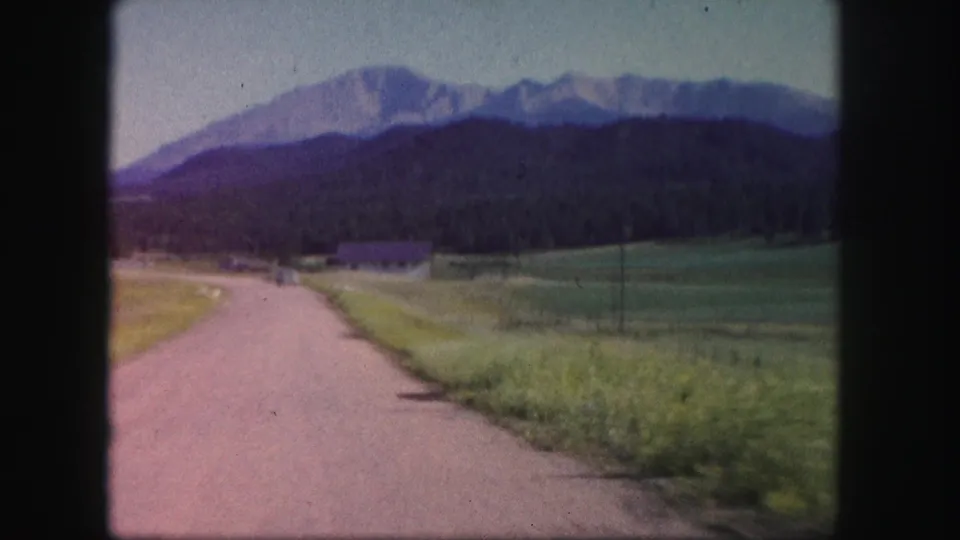 1958 Colorado home-movie frame showing a quiet road through green fields, with a dark tree line and mountain range beyond.