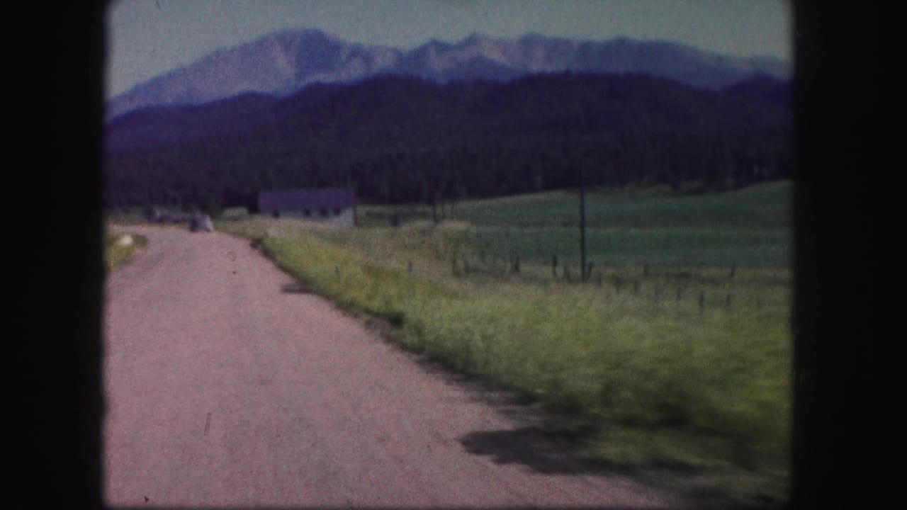1958 Colorado 8mm frame showing a moving view along a dirt road beside open fields and distant mountain ridges.