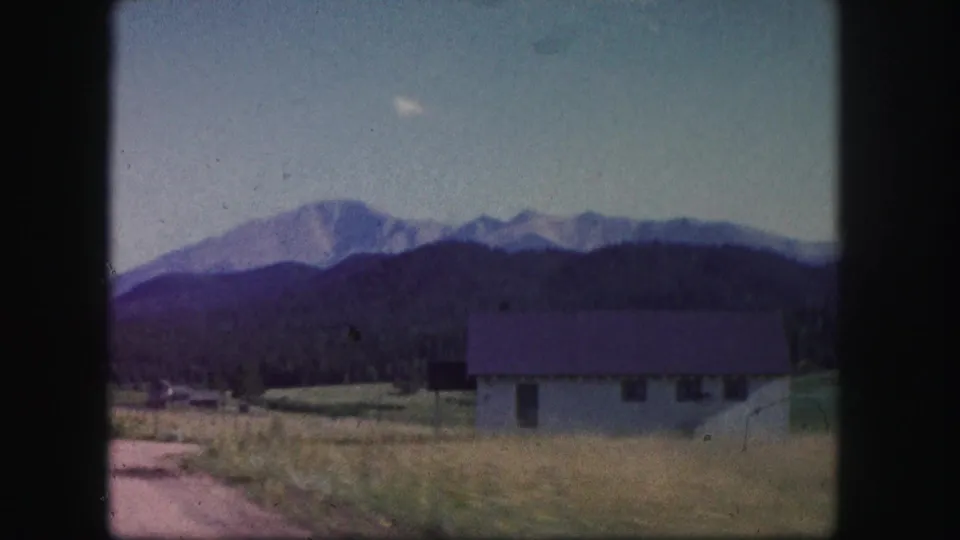 1958 Colorado 8mm frame showing a small house in a grassy field, with dark wooded foothills and mountains beyond.