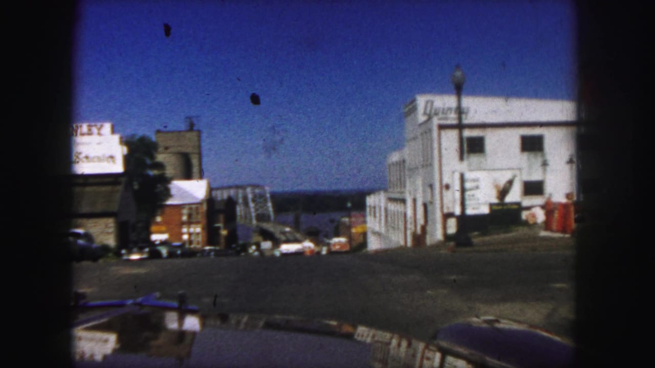 1958 Denver home-movie view from a car approaching a street lined with commercial buildings and period automobiles.