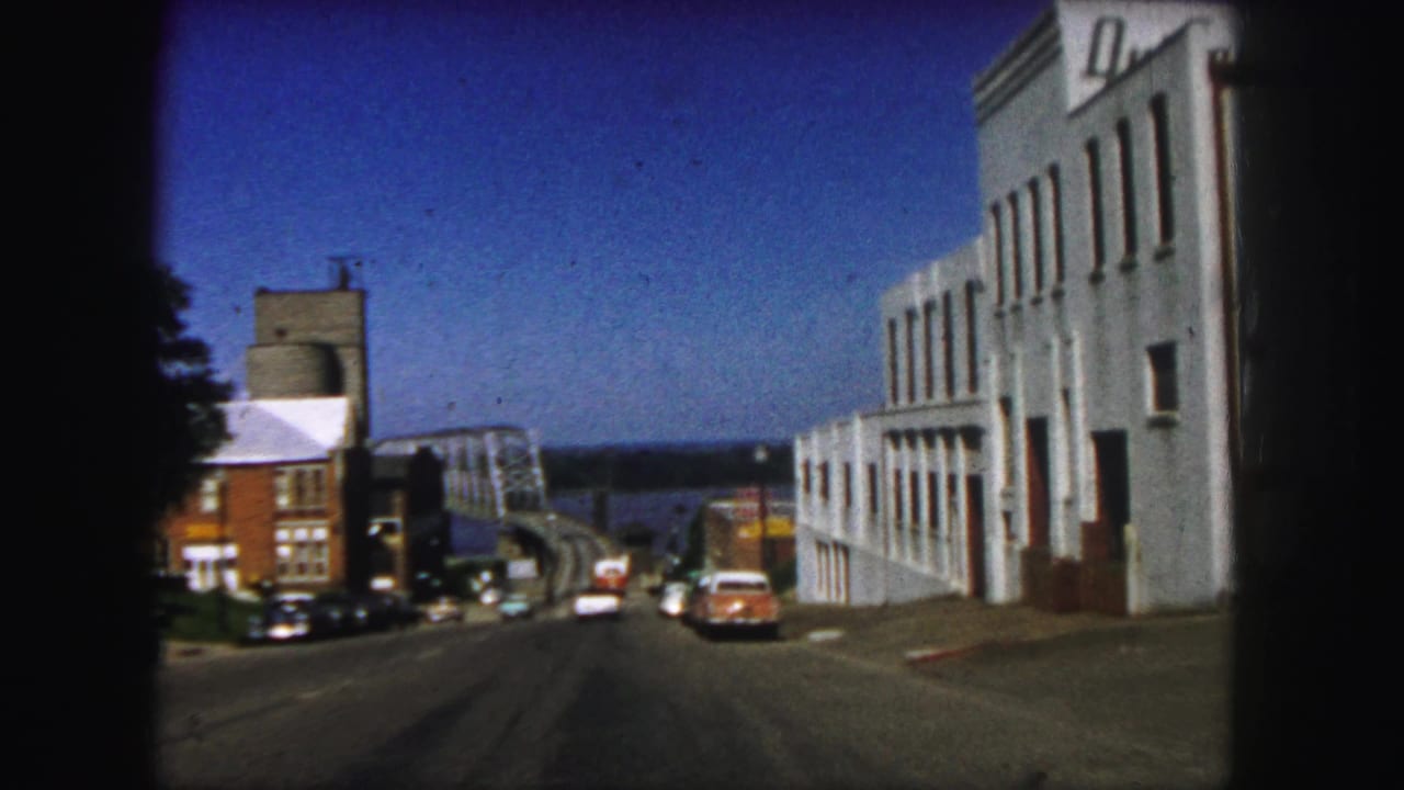 Street approach toward a steel bridge with parked and moving cars beside low-rise buildings.