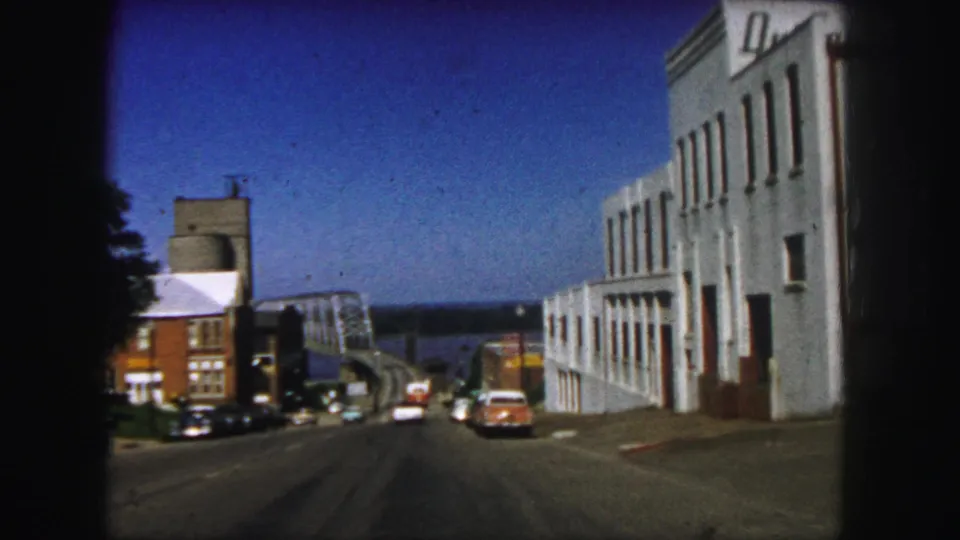 Denver street scene from a moving vehicle, showing mid-century cars, storefronts, and a bridge near the horizon.