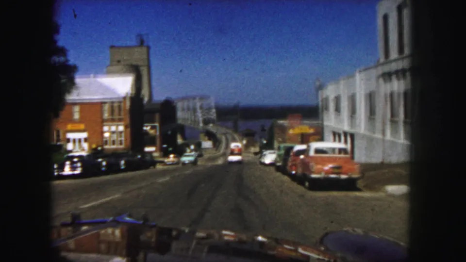 1958 Denver 8mm frame showing a downhill urban road, parked classic cars, and a bridge in the distance.