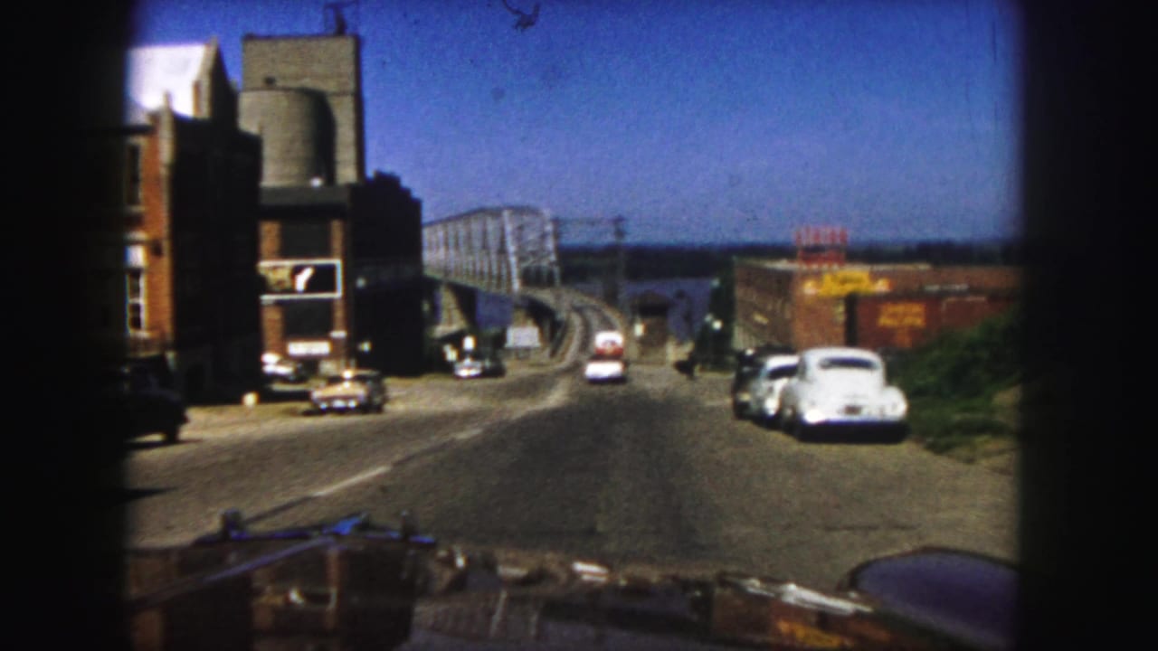 1958 Denver home-movie view from a car descending a road toward a truss bridge over water, with mid-century cars and roadside buildings.