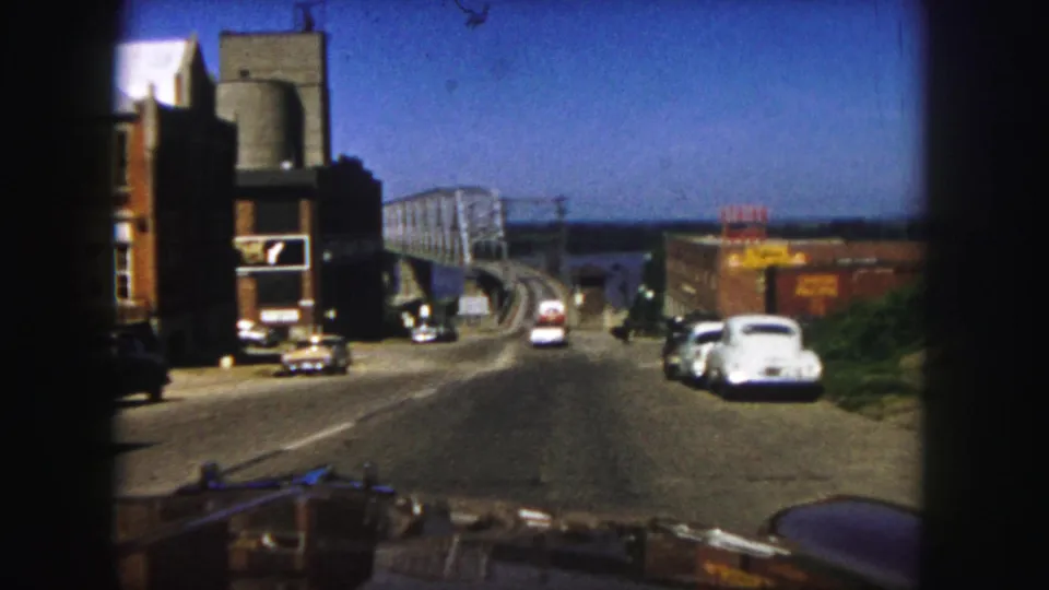 1958 Denver home-movie view from a car descending a road toward a truss bridge over water, with mid-century cars and roadside buildings.