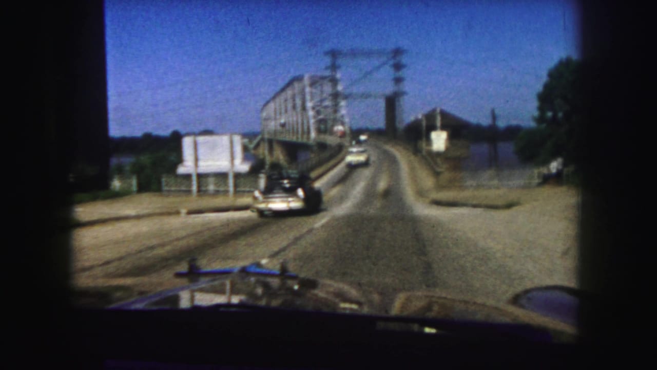 First-person 1958 Denver view nearing a steel truss bridge, with automobiles on the road, utility lines overhead, and water to the side.