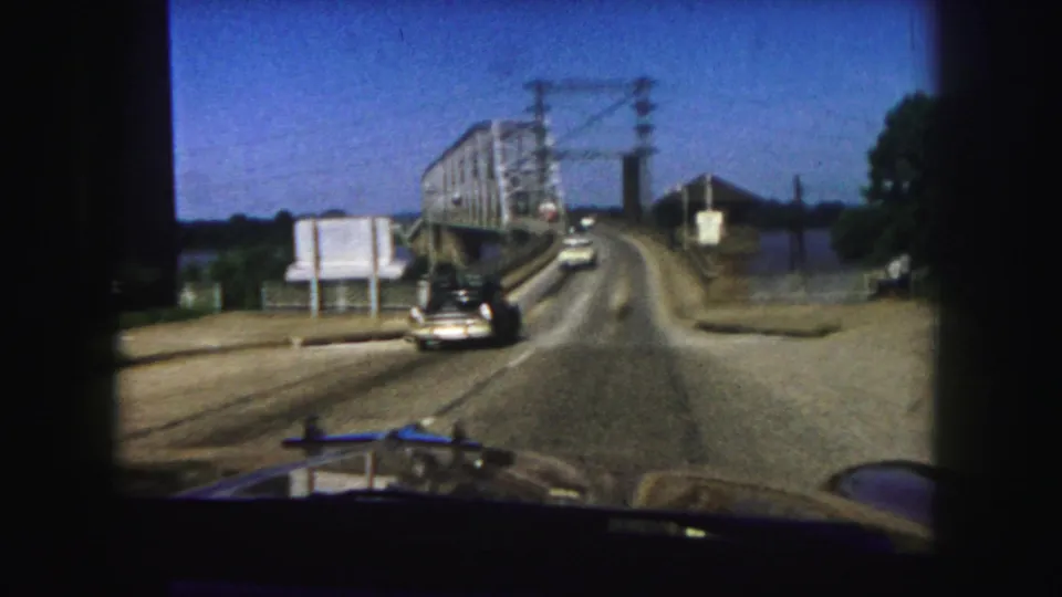 First-person 1958 Denver view nearing a steel truss bridge, with automobiles on the road, utility lines overhead, and water to the side.