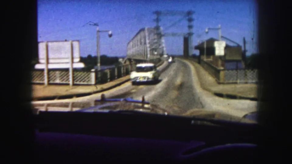 1958 Denver car POV at the bridge approach, showing a vehicle ahead, roadside signs, railings, utility lines, and a small bridge-side structure.