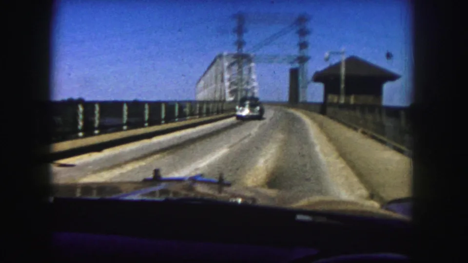 Moving 1958 Denver car view on the bridge deck, facing a steel truss span with railings, overhead utility lines, and water visible beside the roadway.