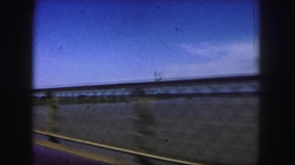 Continuation of the side view across the bridge, showing railing, water, and a bright sky in grainy 8mm color.