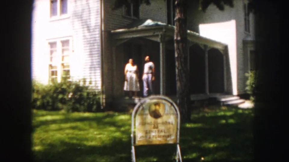 Two adults stand on a porch outside a white clapboard house in the Denver 1958 home-movie footage, with a marker in the foreground.