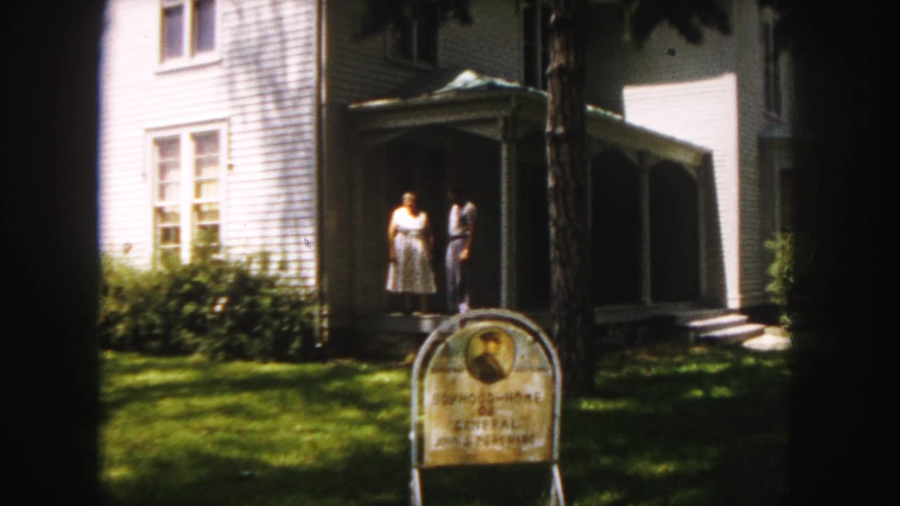 1958 Denver home-movie frame showing an adult couple on the porch of a white clapboard house with a weathered landmark sign on the lawn.