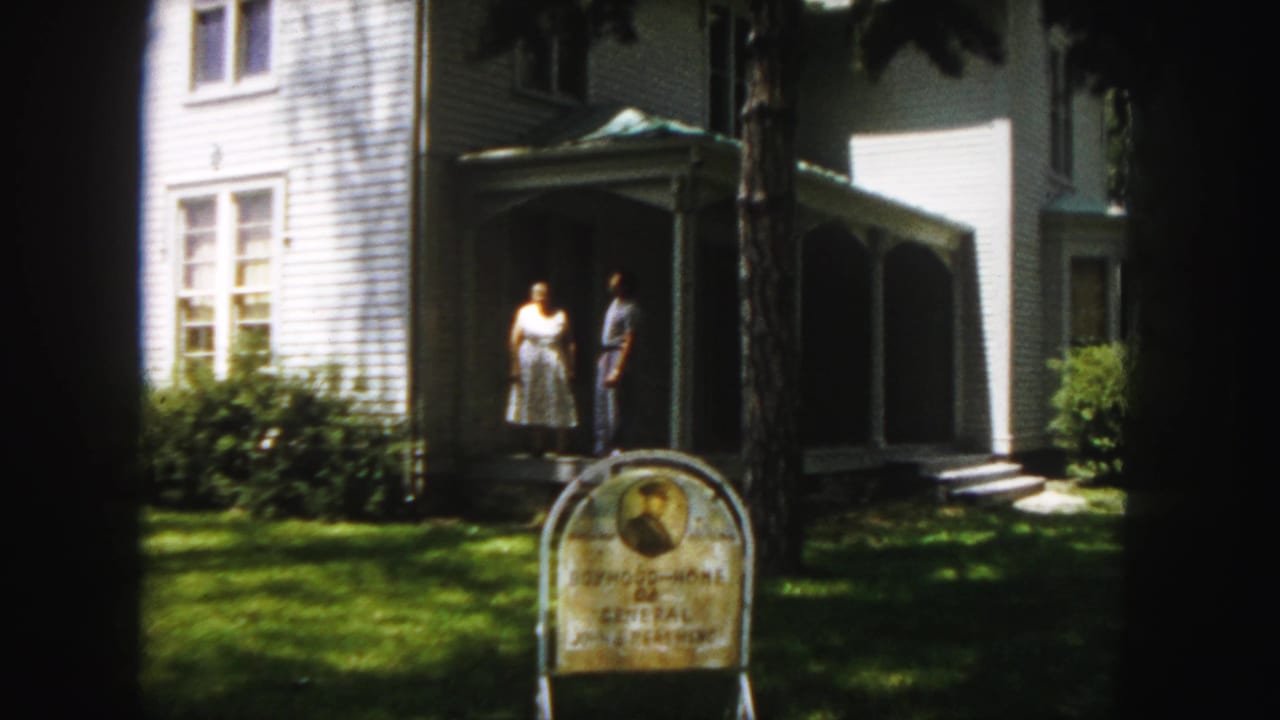 Adult couple stands near the porch entrance with a landmark sign and shaded lawn in front.