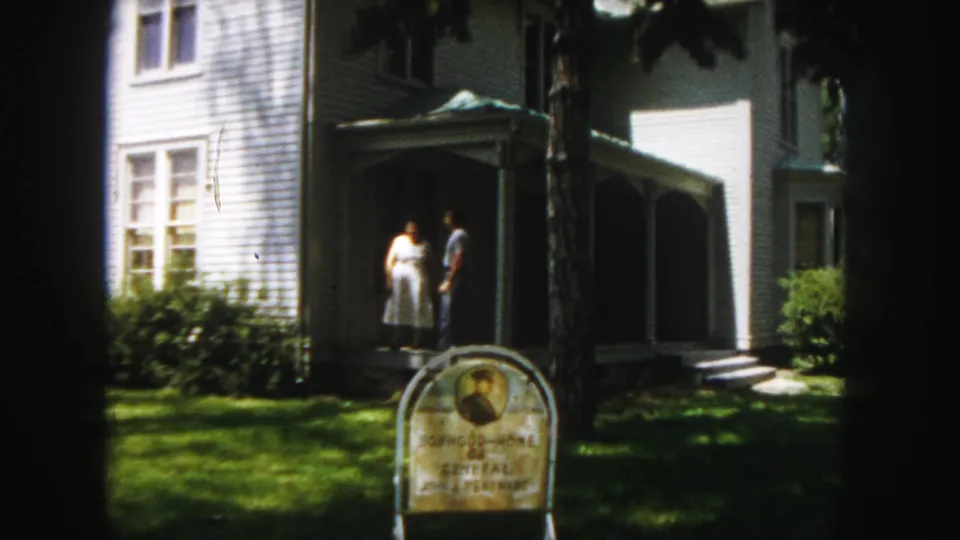 1958 Denver home-movie frame showing the couple closer together near the porch of the white house, with the historic sign partly visible below.