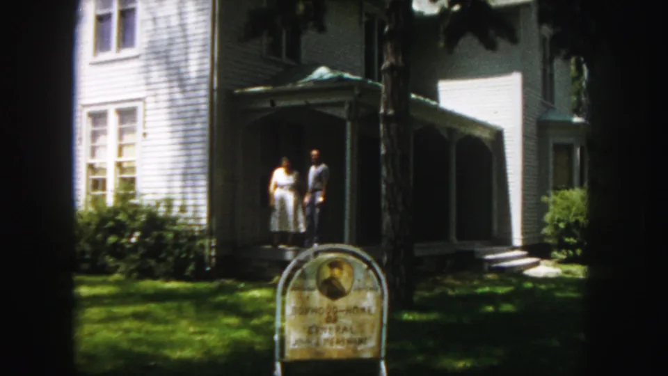 Final 1958 Denver frame holds on the adult couple near the white clapboard house porch, with a foreground landmark marker and sunlit lawn.