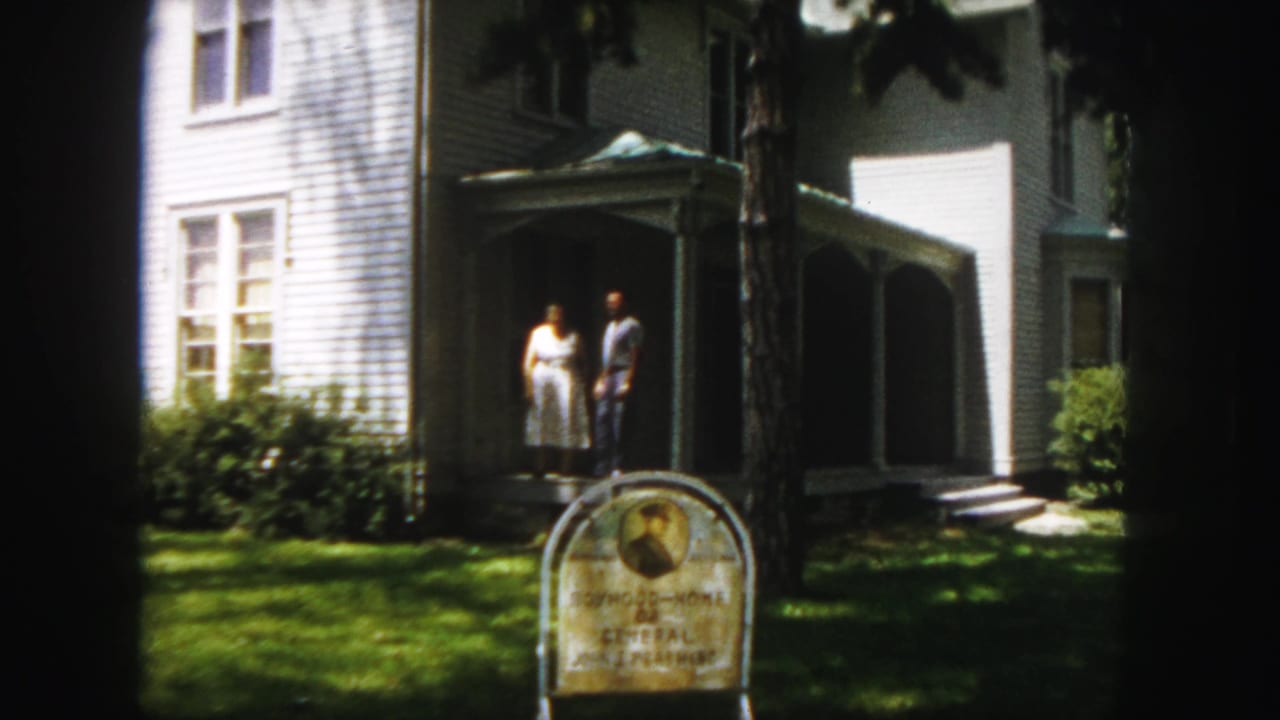 Two adults stand on the porch of a white clapboard house behind a lawn sign.