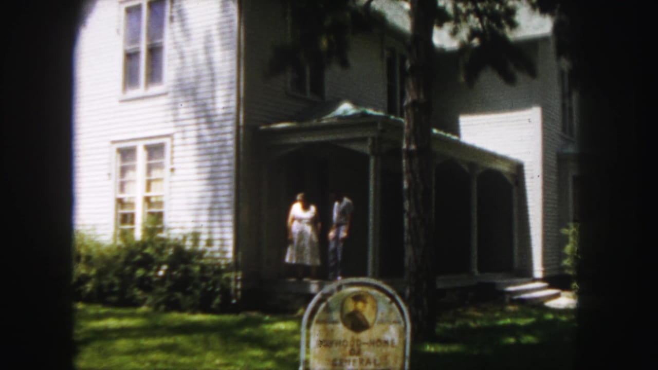 Two adults stand near the porch of a white house with a sign in the foreground.