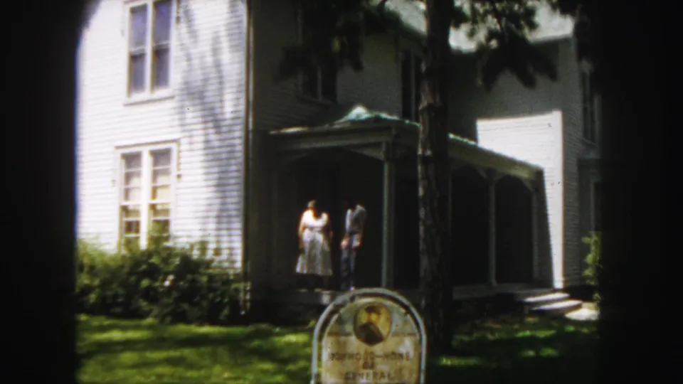 The camera frames the white clapboard house, shaded porch, two adults, and the Boyhood-Home of General John J. Pershing sign.