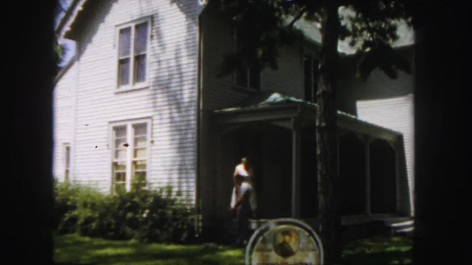 In the 1958 Denver home-movie view, one adult moves near the shaded porch while the historic-site sign remains partly visible on the lawn.