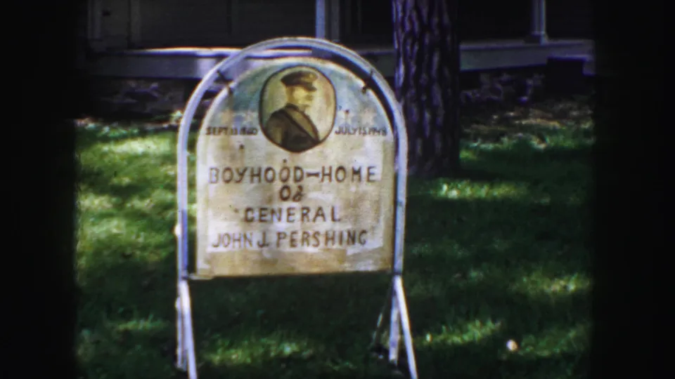 Final 1958 Denver frame showing the Pershing boyhood home sign on white supports, with lawn, tree trunk, and shaded background.