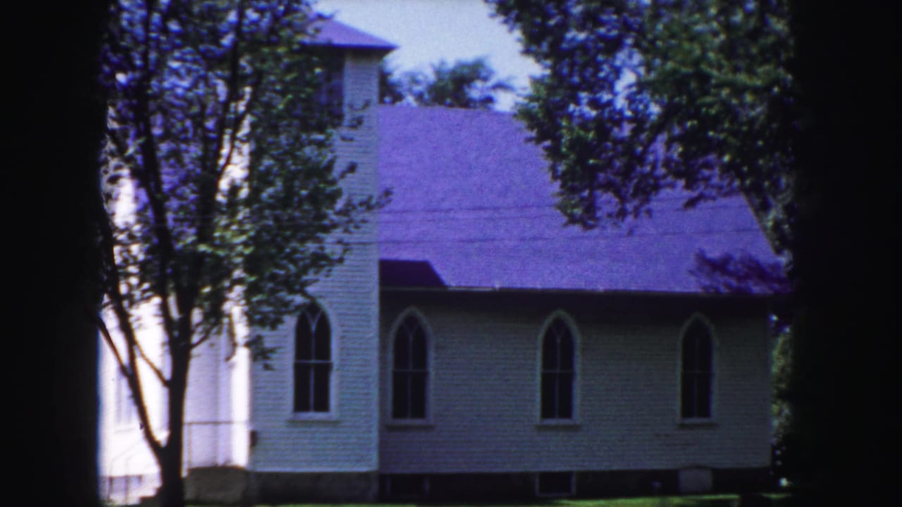 1958 Denver-area home-movie frame of a white church with a purple roof, arched windows, trees, lawn, and dark film-edge shadows.