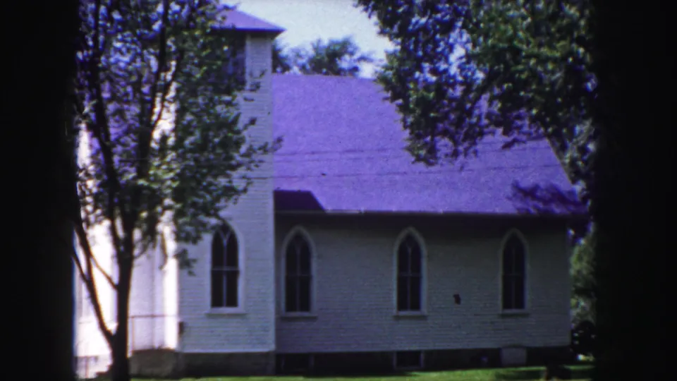 1958 Denver-area 8mm frame showing the church facade partly blocked by tree trunks, with arched windows, roofline, lawn, and dark gate edges.