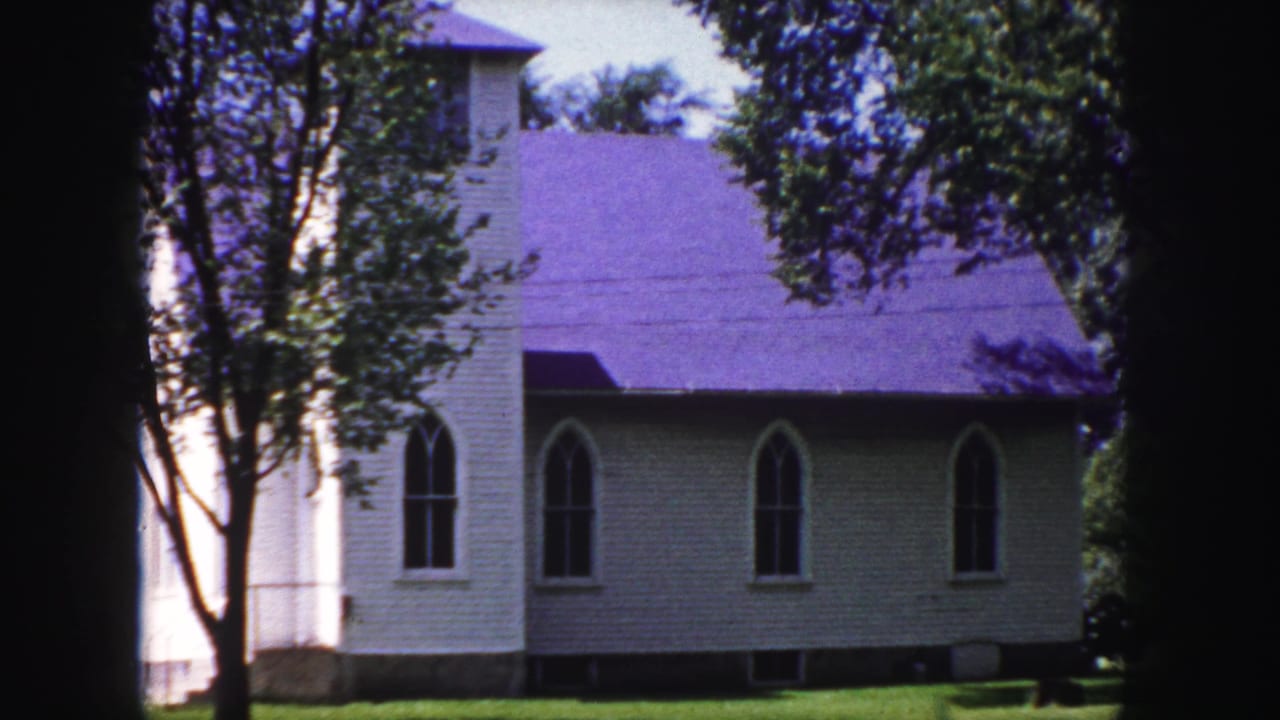 1958 home-movie frame near Denver showing a white Gothic-style church with arched windows, a purple roof, and trees in the foreground.