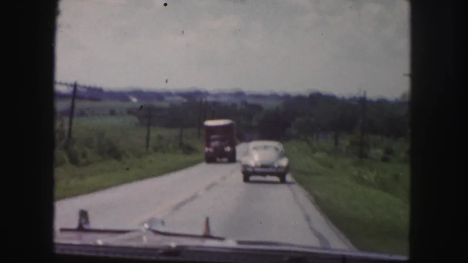 1958 home-movie road view near Denver with a rounded sedan following a truck on a two-lane road through open green countryside.