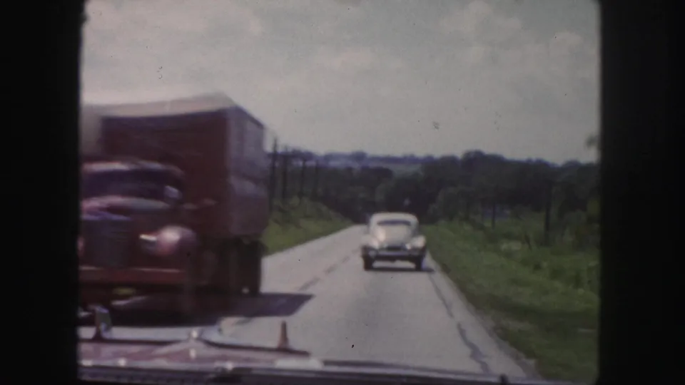 1958 road-travel frame near Denver showing a truck passing close to the camera while a classic sedan drives ahead on a rural two-lane road.