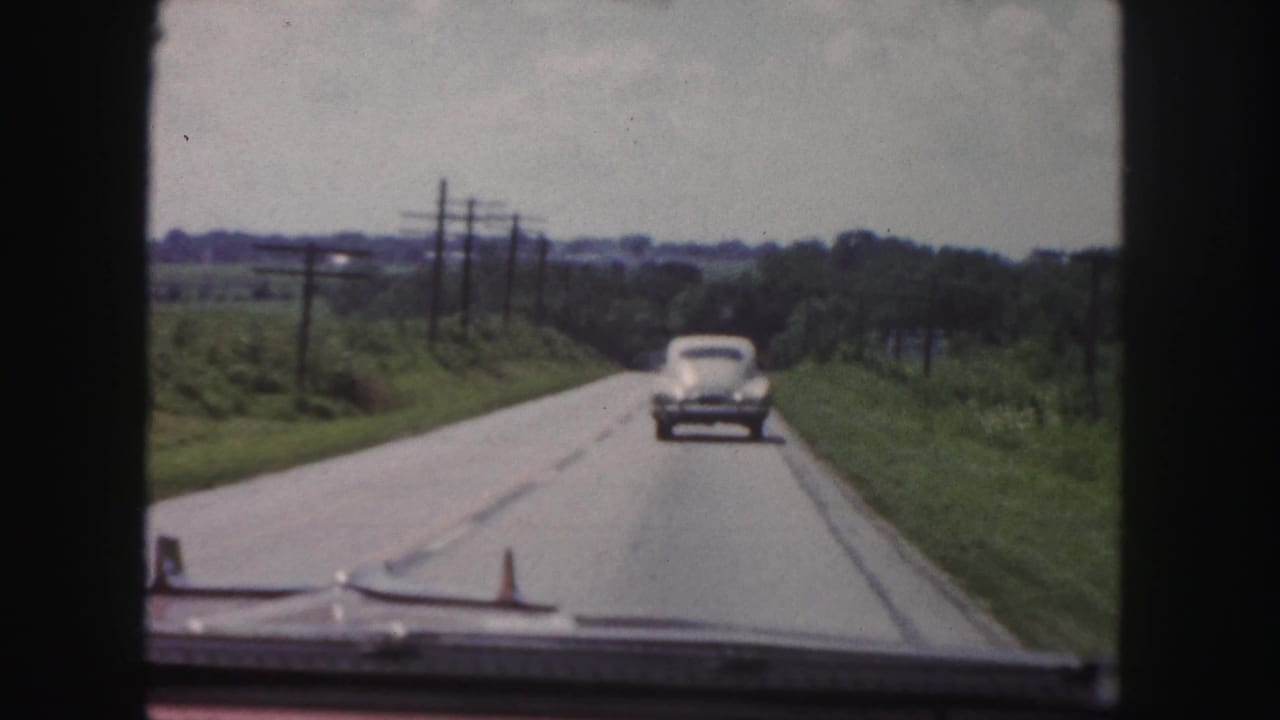 1958 home-movie view from inside a moving car following a rounded sedan along a rural road with fields and utility poles.