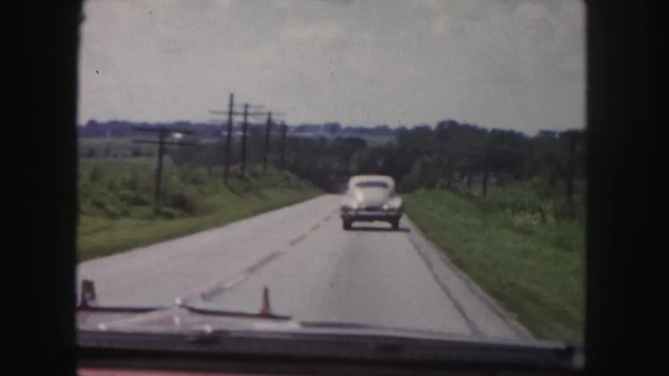 1958 home-movie view from inside a moving car following a rounded sedan along a rural road with fields and utility poles.