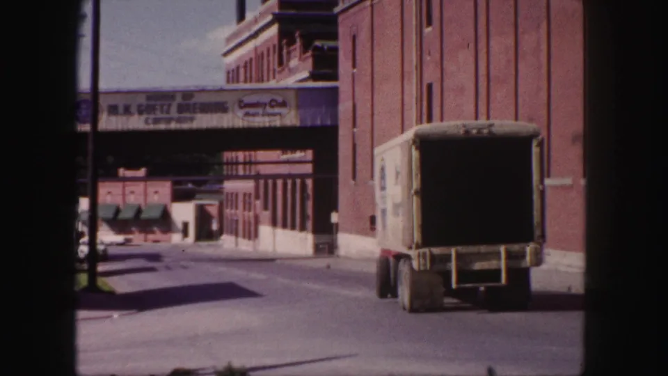Rear view of a delivery truck moving along a Denver industrial street beside brick buildings and brewery-related signs.