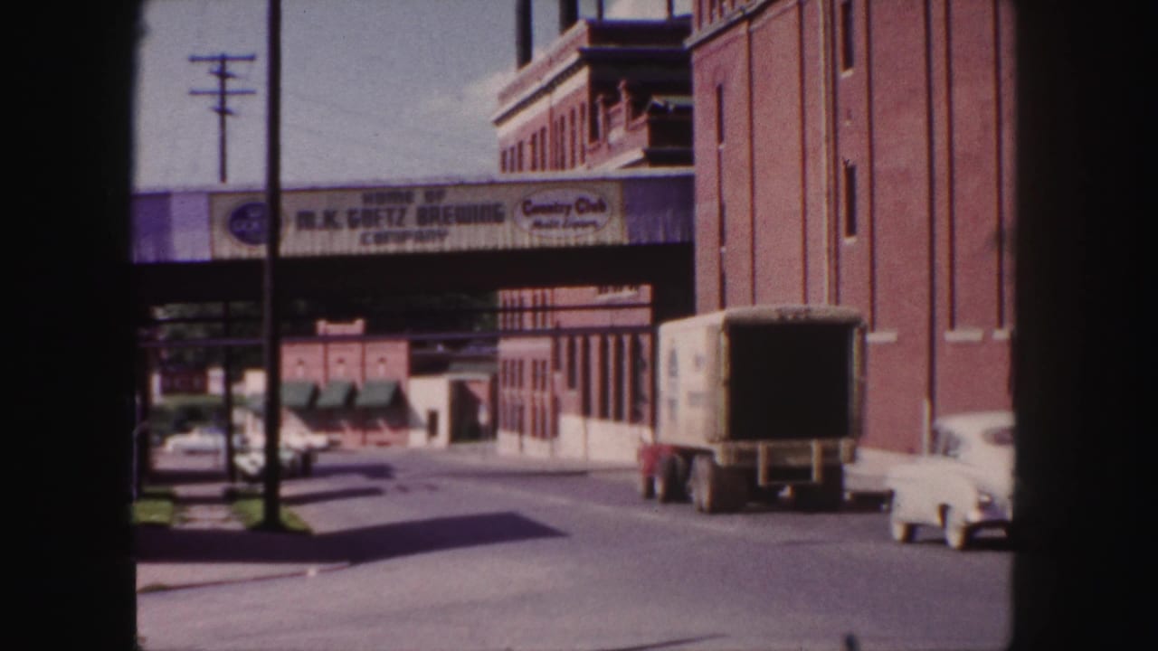 White car and box truck on a sunlit street beneath a Goetz Brewing overpass sign.