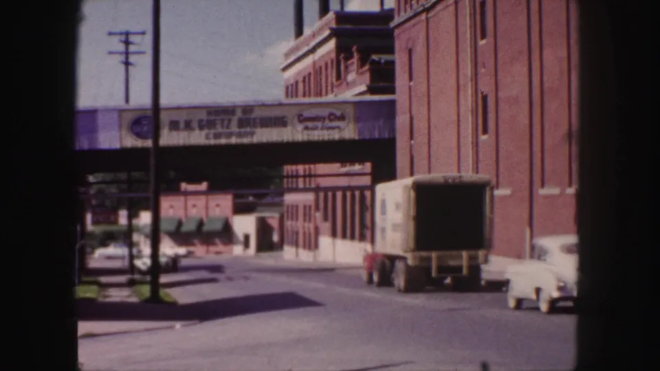 1958 Denver street view with a white car, box truck, brick industrial buildings, and Goetz Brewing overpass signage.