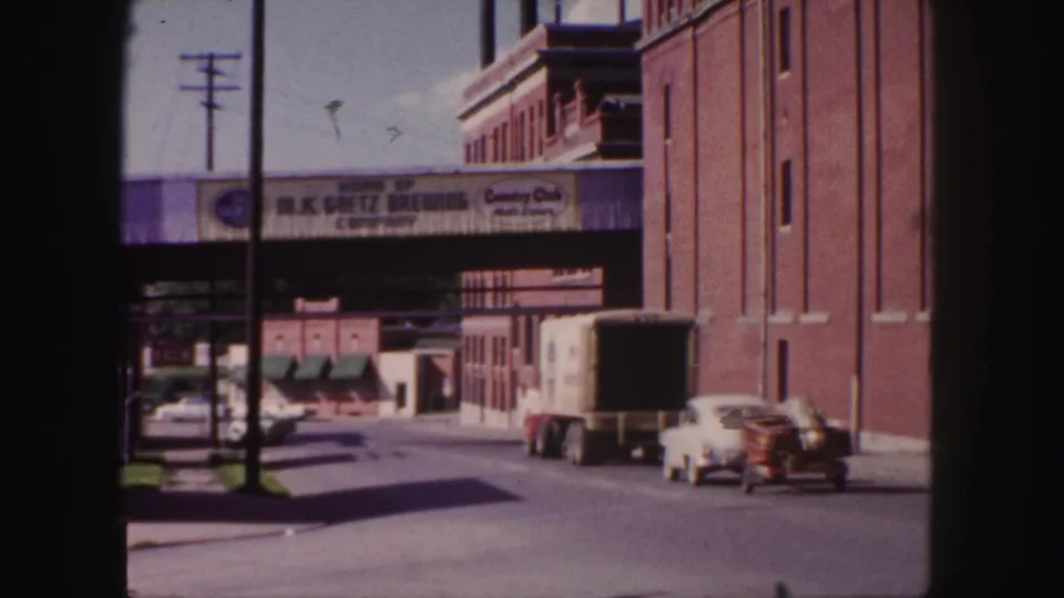 1958 Denver industrial street with truck, white car, parked vehicles, and visible Goetz Brewing advertising over the roadway.