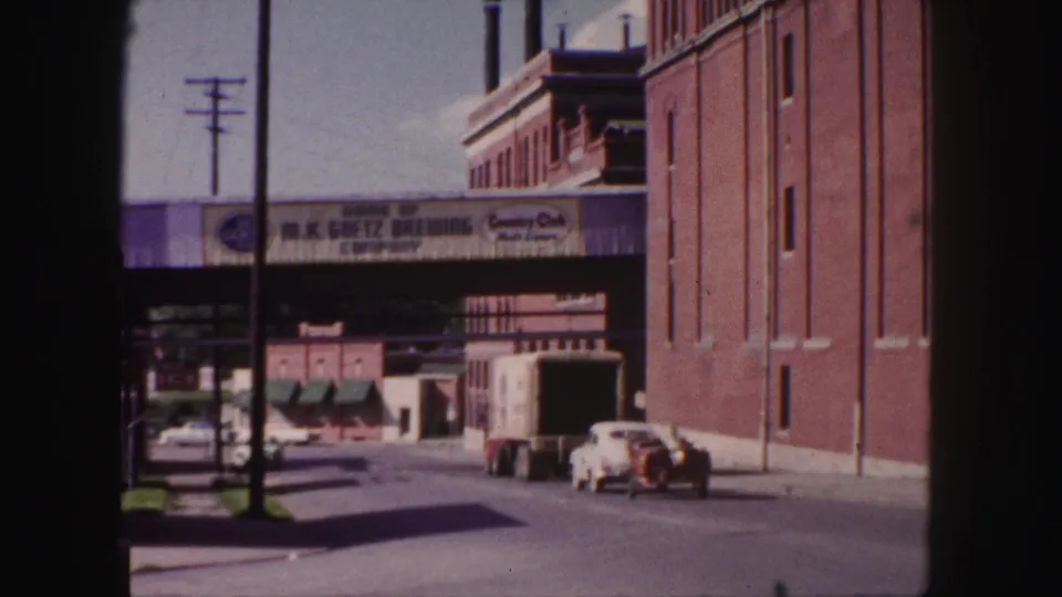 1958 Denver street scene showing a box truck, white car, brick industrial facades, and brewery advertising on an overpass.