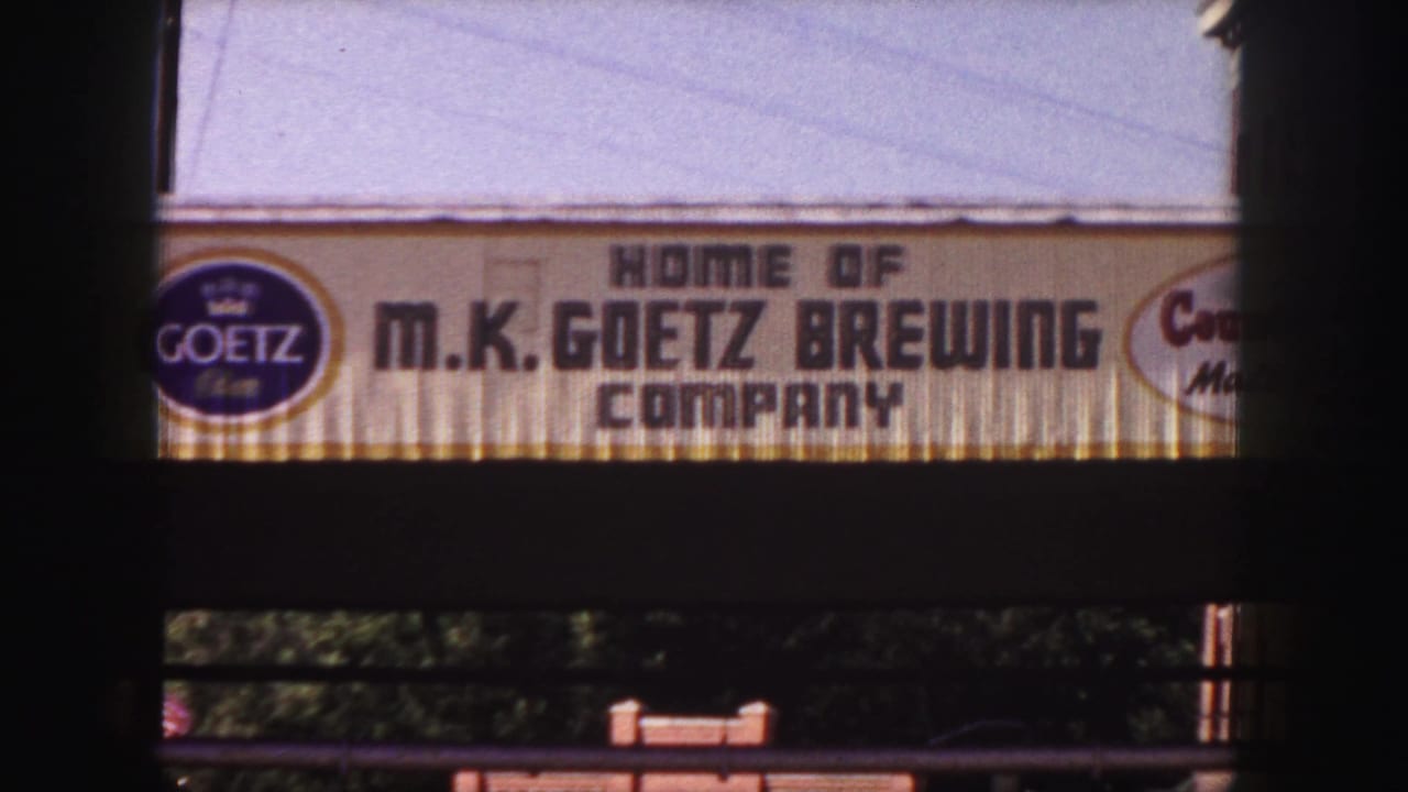 1958 Denver home-movie frame centered on the M.K. Goetz Brewing Company sign beneath a pale sky.