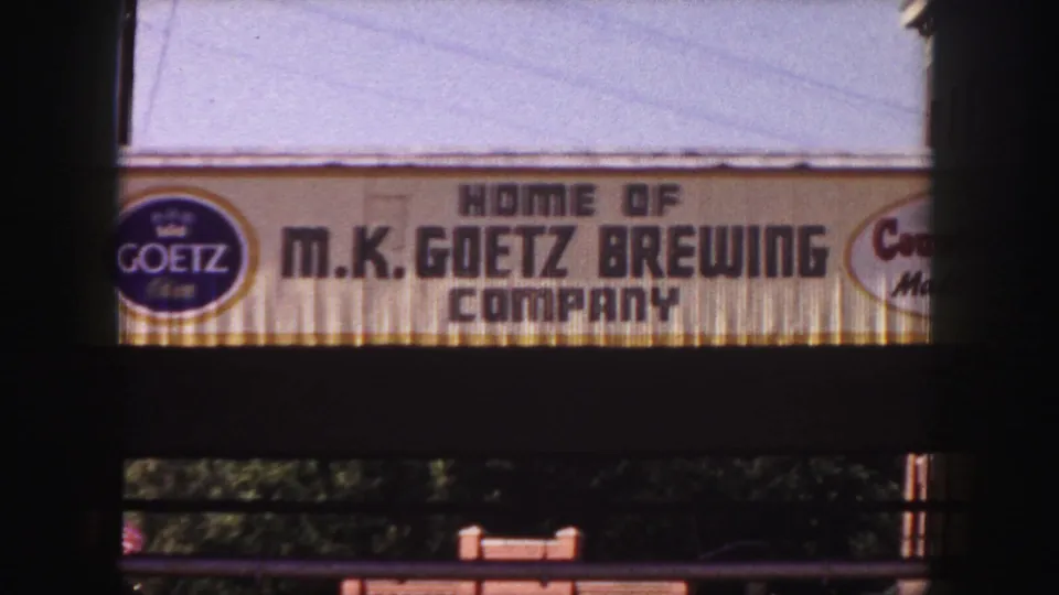 1958 Denver home-movie frame centered on the M.K. Goetz Brewing Company sign beneath a pale sky.