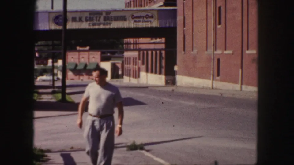 1958 Denver 8mm frame showing an adult man walking toward the camera near the brick M.K. Goetz Brewing Company exterior.