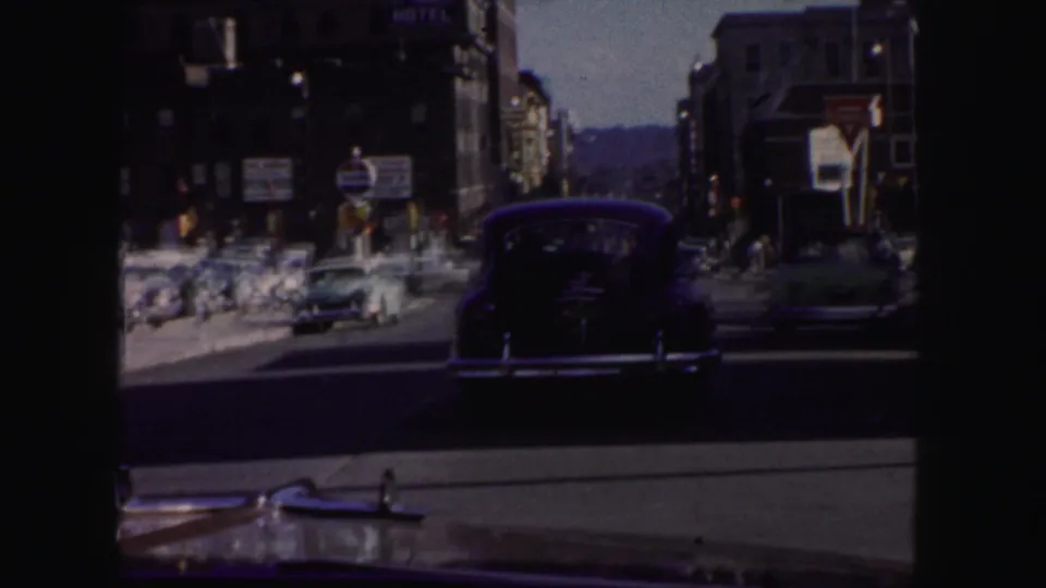 Denver street scene with mid-century cars, storefront signs, and the hood of a vehicle in the foreground.