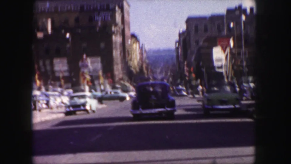 1958 Denver street view with vintage cars, tall buildings, and a blurred cyclist or pedestrian near the roadway.