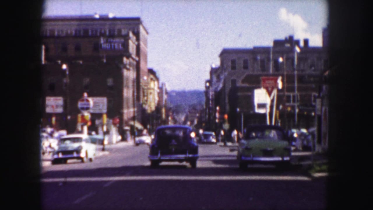 1958 Denver street view with mid-century cars moving through a broad urban road beneath brick buildings and traffic signage.