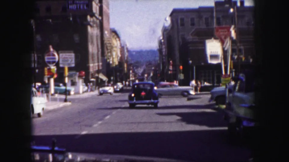 1958 Denver downtown road scene with a dark car centered in the lane, parked cars at the sides, and street signage overhead.