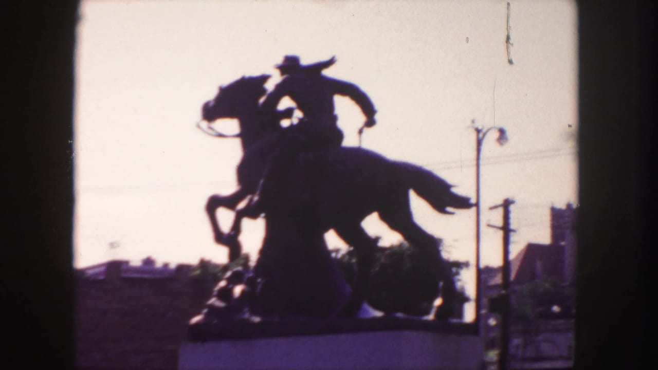 Silhouetted cowboy on horseback statue against a bright sky with urban poles and buildings behind it.