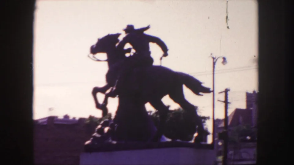 1958 Denver home-movie frame showing a dark equestrian cowboy statue on a pedestal, backlit against the sky with street fixtures nearby.