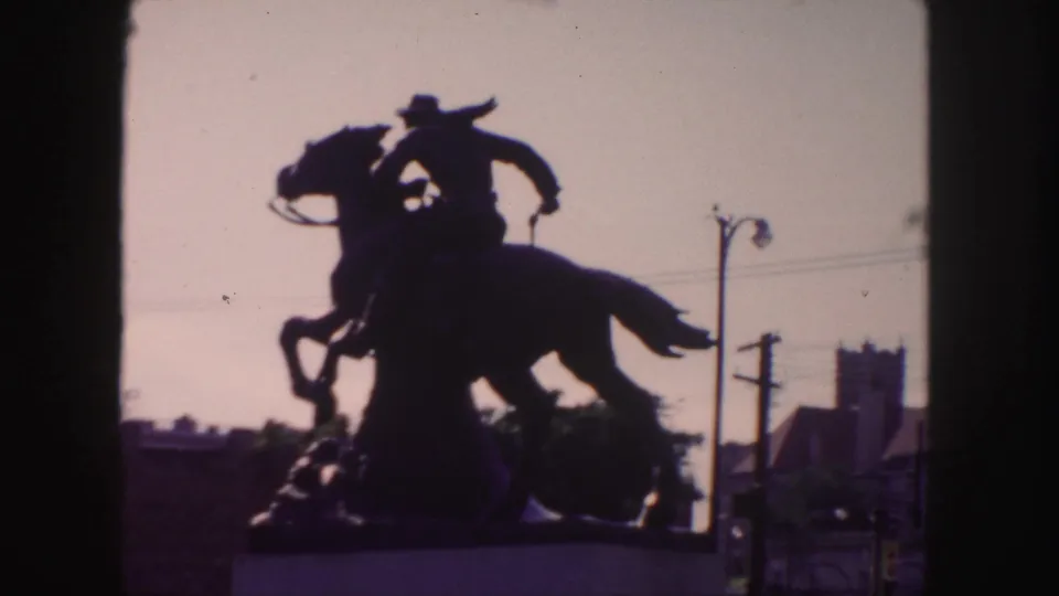 1958 Denver frame of a silhouetted mounted cowboy statue, with the raised horse form set against sky and urban street details.
