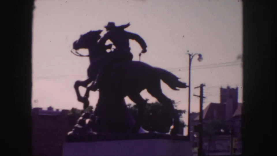1958 Denver home-movie still of a backlit equestrian cowboy statue on a pedestal, surrounded by visible urban street elements.